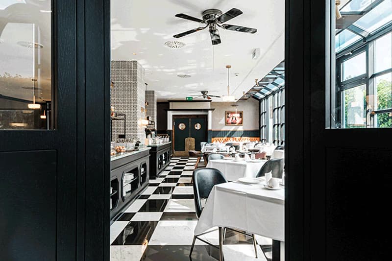 Restaurant mit schwarz-weiss kariertem Boden, gedeckten Tischen, Buffetzeile links und grossen Fenstern rechts.