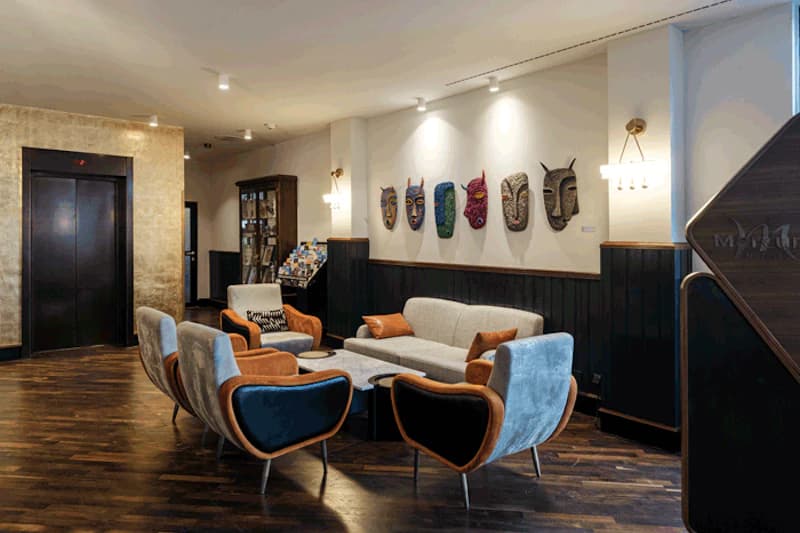 Hotel-Lounge mit Sofas und Sesseln, Wand mit fünf dekorativen Masken, Broschürenständer und Aufzugstür.