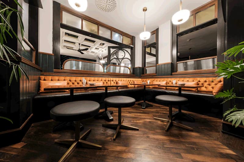 Eckbank mit gestepptem cognacfarbenem Leder, runde schwarze Hocker, Holzboden und hängende Lampen in einer Hotelbar.