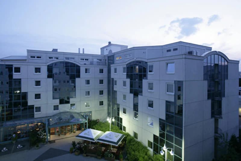Mehrstöckiges Hotel mit Glasfassaden, überdachtem Eingang und Außenbestuhlung unter Sonnenschirmen.