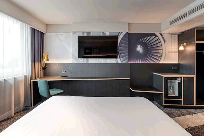 Hotelzimmer mit Bett im Vordergrund, Wandfernseher, Schreibtisch mit Stuhl, Minibar und Turbinenbild an der Wand.