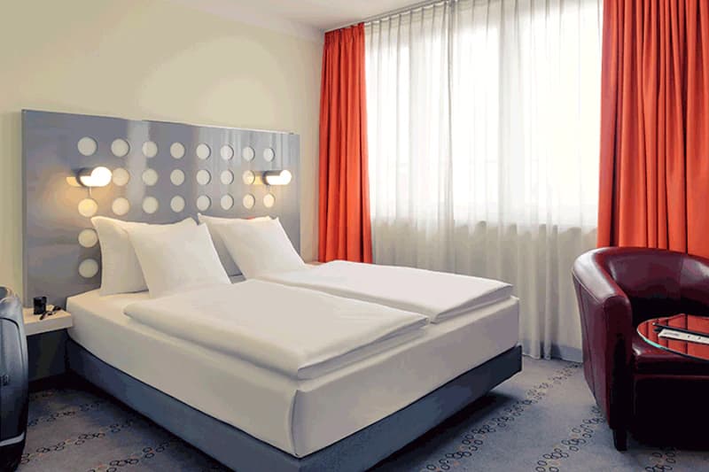Hotelzimmer mit zwei Einzelbetten, grauem Kopfteil mit runden Aussparungen, orangefarbenen Vorhängen und rotem Sessel.