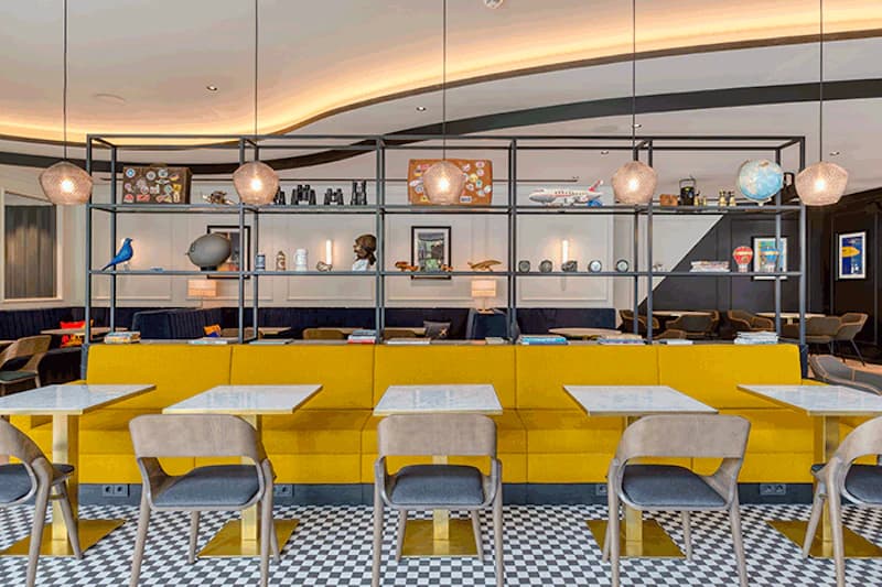 Hotel Lounge mit gelber Sitzbank, Tischen, Stühlen und offenen Regalen mit Dekorationen.