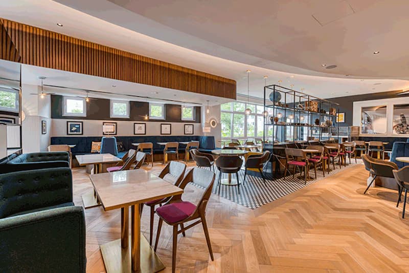 Heller Hotelgastraum mit Tischen, Stühlen, Sitzbänken, großen Fenstern und hölzernem Fischgrätboden.