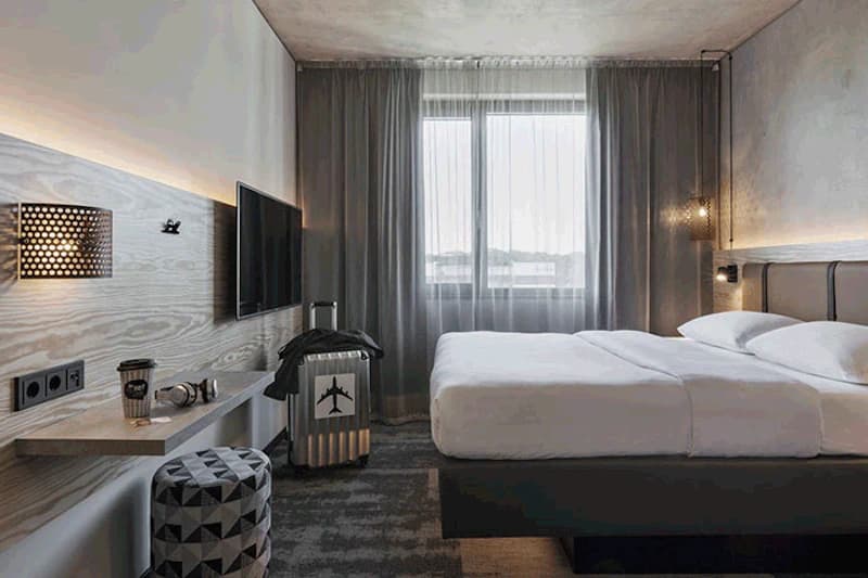 Hotelzimmer mit Doppelbett, Fenster mit Gardinen, Wandfernseher, Reisekoffer mit Flugzeugsymbol und Ablagetisch.