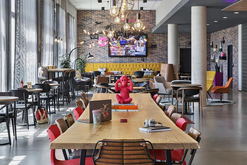 Große Hotellounge mit langem Holztisch, roten Stühlen, roter Affenskulptur, gelber Sitzbank und Wand mit Fernseher.
