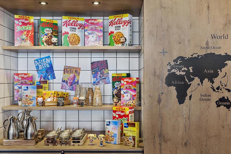 Regal mit bunten Kelloggs-Müslischachteln, Glasbehältern, Kaffeekannen und Weltkarte an Holzwand.
