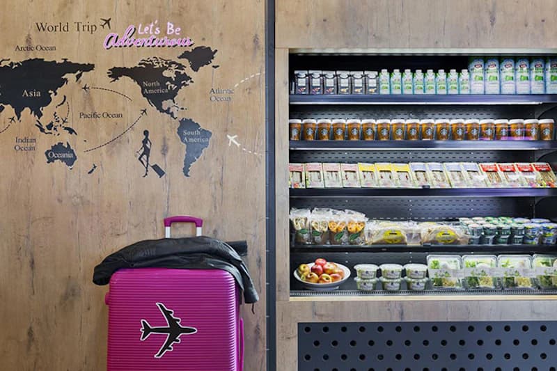 Pinkfarbener Koffer mit Flugzeugaufkleber vor Holzwand mit Weltkarte und Schriftzug Let's Be Adventurous, daneben gekühltes Regal mit Snacks.
