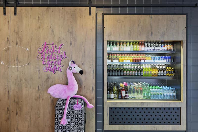 Holzwand mit rosa Stoffflamingo auf einem Koffer neben beleuchtetem Getränkekühlschrank.