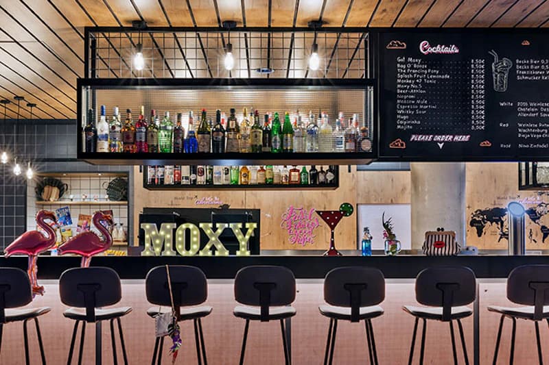 Moderne Hotelbar mit Tresen, fünf Hockern, beleuchtetem MOXY-Schriftzug, Regal mit Spirituosen und hängenden Lampen.