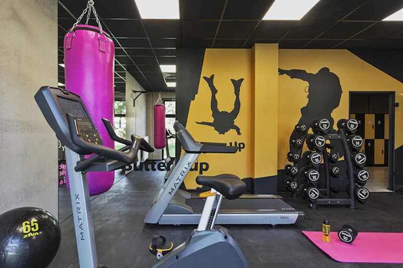 Fitnessraum mit Laufband, Ergometer, rosa Boxsäcken, Hanteln auf Ständer und Wandgrafiken in Gelb und Schwarz.