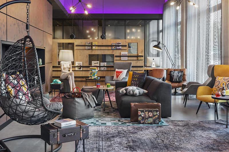 Hotel-Lounge mit Hängesessel, Sofas, Regalen, Stehlampe und großen Fenstern mit Vorhängen, violette Deckenbeleuchtung.