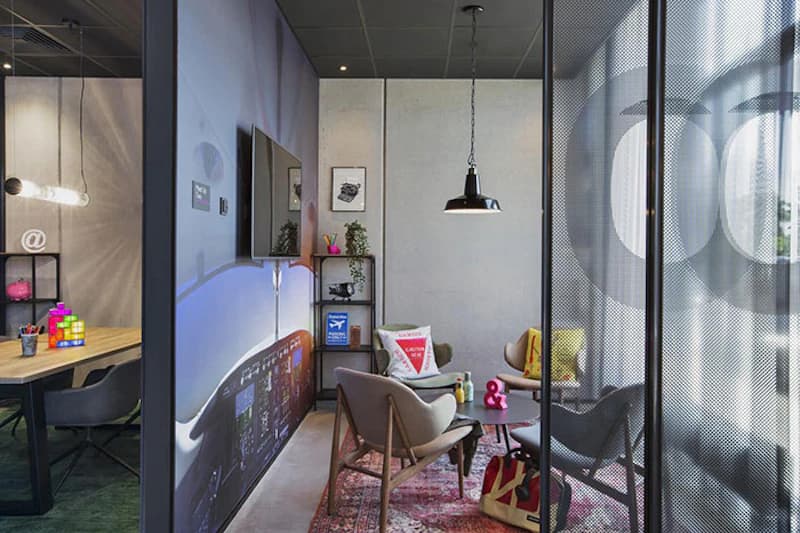 Lounge mit Sesseln, kleinem Tisch, Hängelampe, Regal und Wandgrafik eines Cockpits, getrennt durch gelochte Metallwand.