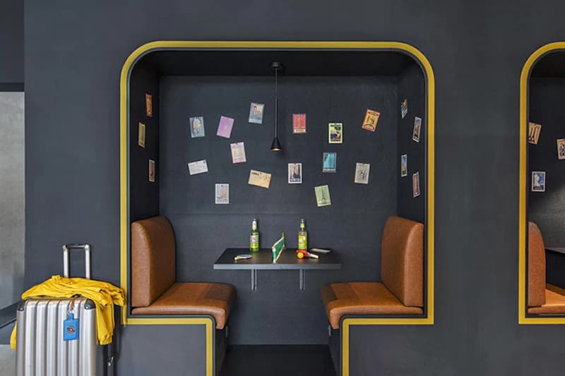 Zwei Sitznischen mit braunen Polstersitzen, kleinem Tisch, zwei Flaschen und Postkarten an dunkler Wand; links Rollkoffer mit gelber Jacke.