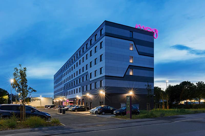 Mehrstöckiges Hotelgebäude mit beleuchtetem moxy-Schriftzug und geparkten Autos bei Abenddämmerung.