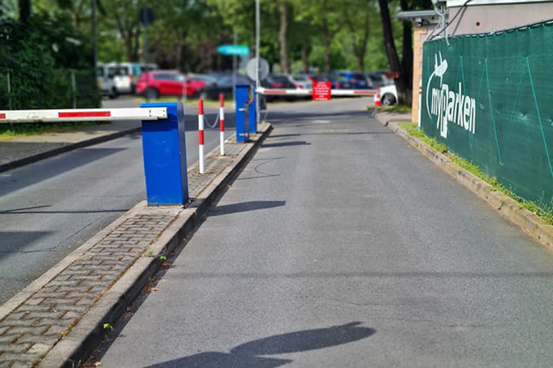 Zufahrt zu Parkplatz mit zwei Schranken, blauen Automaten, roten Leitpfosten und grünem myparken-Zaun.