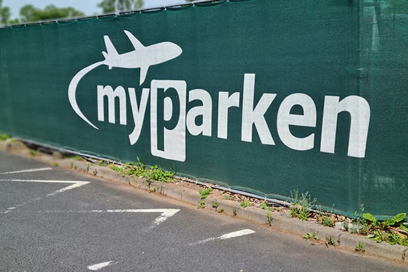 Grüne Zaunplane mit weißem myparken-Logo und Flugzeugsymbol vor markierten Parkflächen.