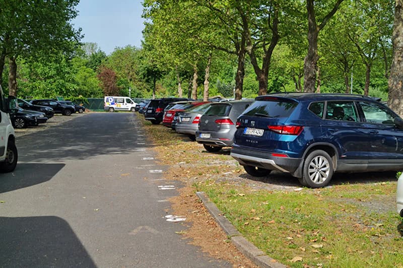 Reihenparkplatz mit geparkten Autos rechts neben einer Fahrspur, Bäume im Schatten und Lieferwagen vor einem Zaun im Hintergrund.
