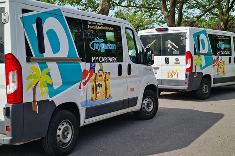 Zwei weiße Shuttlebusse mit myparken-Logo und Kofferaufklebern auf einem Parkplatz unter Bäumen.