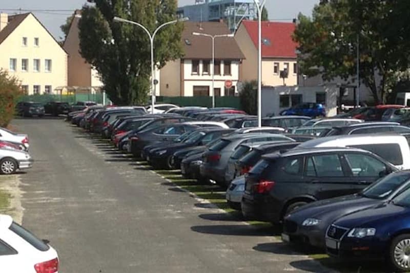 Großer Außenparkplatz mit zwei Reihen geparkter Autos, Straßenlampen und Wohnhäusern im Hintergrund.