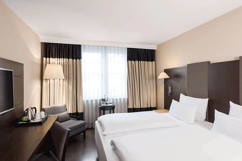 Hotelzimmer mit zwei Betten, Nachttischlampen, Sessel, Schreibtisch mit Wasserkocher und Fenster mit Vorhängen.