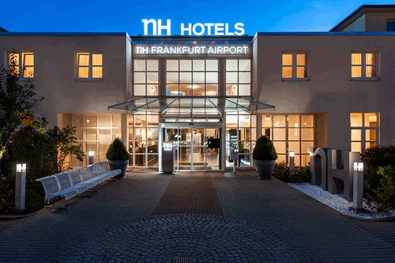 Beleuchteter Hoteleingang mit Glasvordach, Sitzbank und leuchtendem NH FRANKFURT AIRPORT Schriftzug.