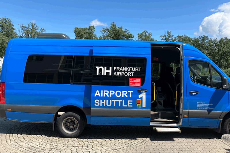 Blauer Kleinbus mit NH Frankfurt Airport Schriftzug und offener Seitentür auf gepflastertem Vorplatz.