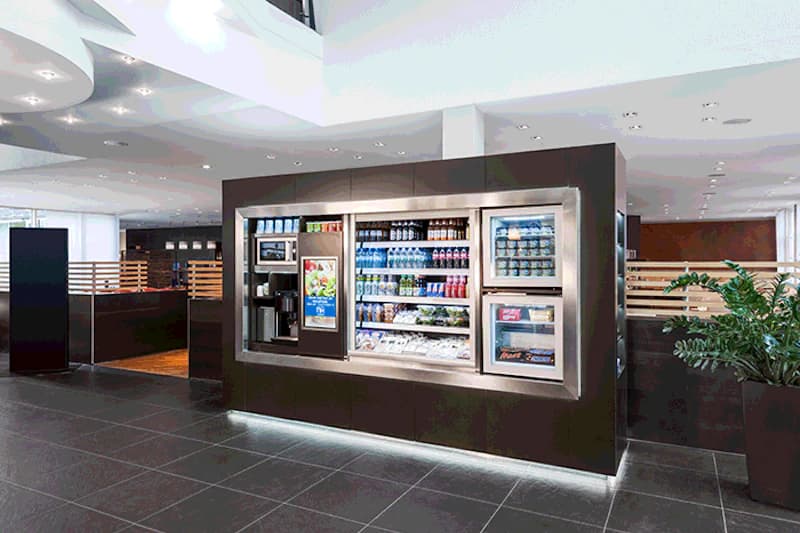 Selbstbedienungsbereich mit Kühlschrankvitrine, Getränken, Snacks und Kaffeemaschine in modernem Hotelfoyer.
