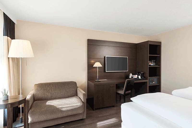 Hotelzimmer mit zwei Betten, Sofa, Schreibtisch, Wandfernseher und Stehlampe.