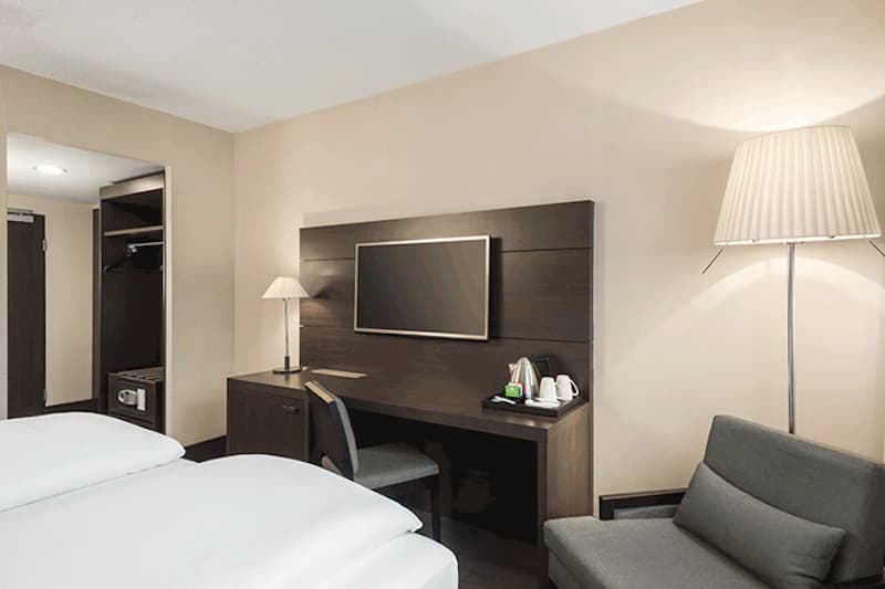 Hotelzimmer mit zwei Betten, Schreibtisch mit Fernseher, Teestation, Sessel und geöffneter Garderobe mit Safe.