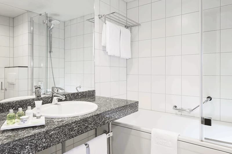 Weisses gefliestes Badezimmer mit Waschbecken, grauer Granitablage, Badewanne, Duschwand und Handtuechern.