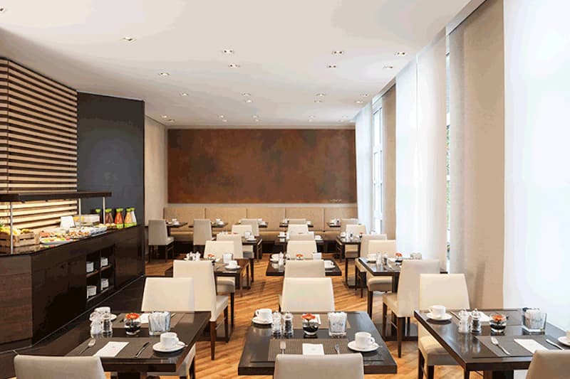 Heller Frühstücksraum mit gedeckten Tischen, weißen Stühlen, Buffet links und großen Fenstern rechts.