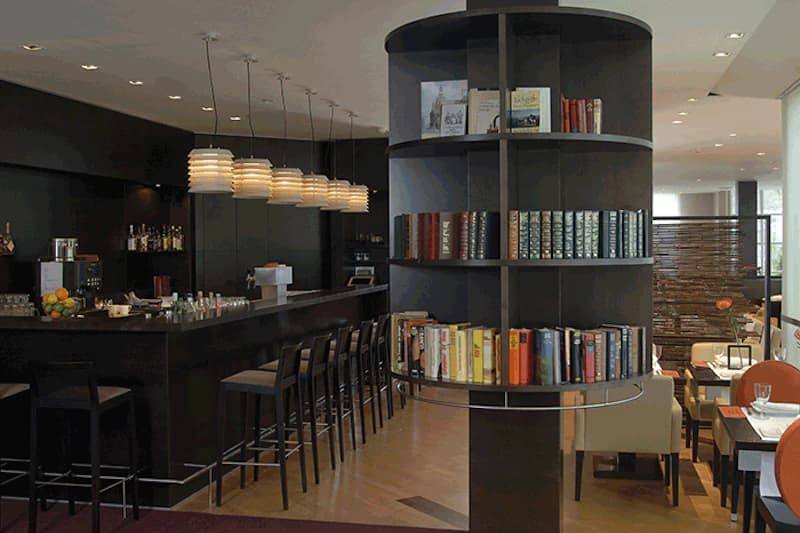 Runde Bücherregal-Säule vor Bartresen mit Hockern, Pendelleuchten und Sitzbereich im Hotel.
