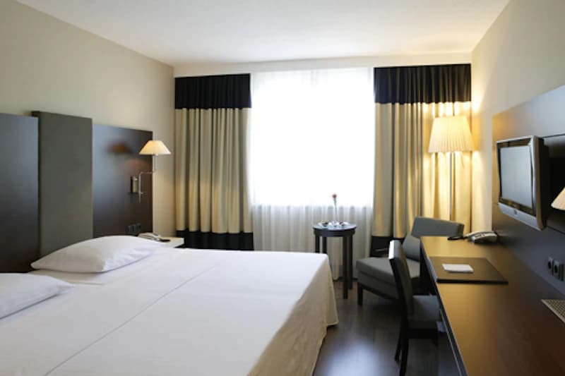 Hotelzimmer mit Doppelbett, großes Fenster mit Vorhängen, Schreibtisch, Sessel und Wandfernseher.