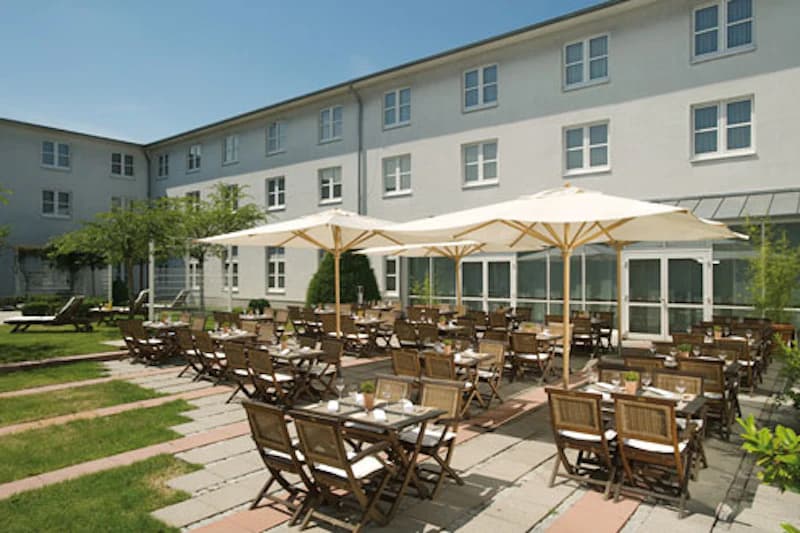Hotelterrasse mit Holzstühlen und Tischen unter weißen Sonnenschirmen vor mehrstöckiger Hotelfassade.