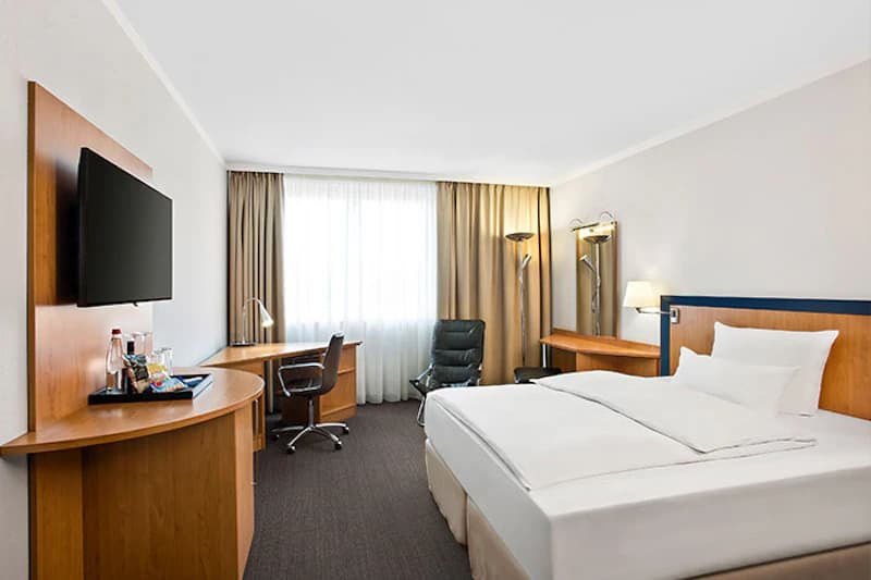 Hotelzimmer mit Doppelbett, Schreibtisch, Wandfernseher, schwarzem Sessel und großem Fenster mit Vorhängen.