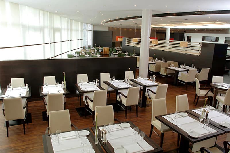 Gedeckte Hotelgastronomie mit Holzboden, cremefarbenen Stühlen und großen Fensterfronten.