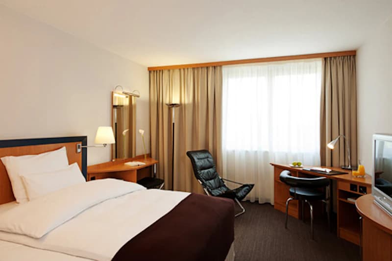 Hotelzimmer mit Doppelbett, Fenster mit Vorhängen, Schreibtisch, Sessel und Fernseher.