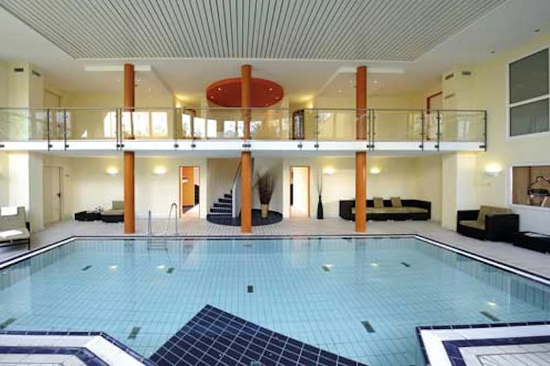 Hallenbad mit hellem Becken, zweigeschossiger Galerie, Glasgeländer und orangefarbenen Säulen.