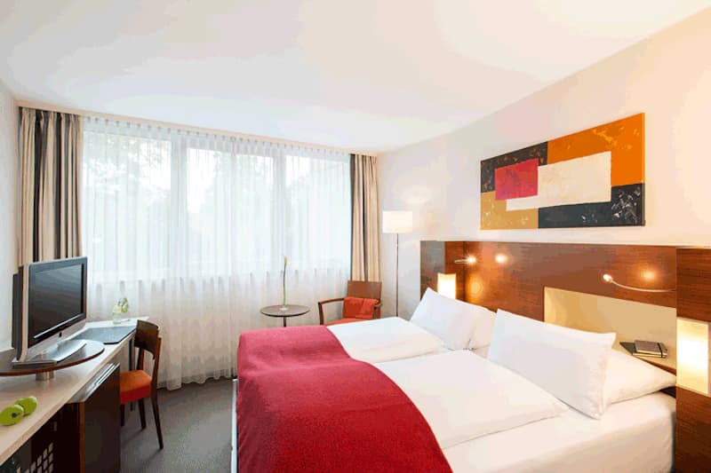 Hotelzimmer mit zusammengestellten Betten, rotem Überwurf, großem Fenster, Schreibtisch mit Fernseher und Sitzbereich.