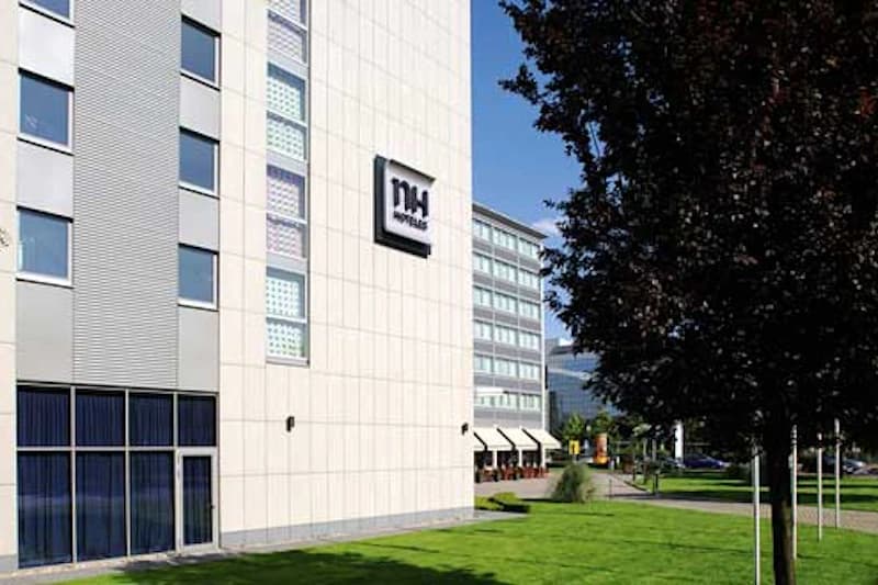Fassadenansicht des NH Hotels mit Logo, Rasenfläche und Baum vor dem Gebäude.