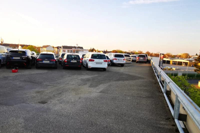Dachparkdeck mit mehreren geparkten Autos und seitlichem Geländer, Häuser am Horizont bei Abendlicht.