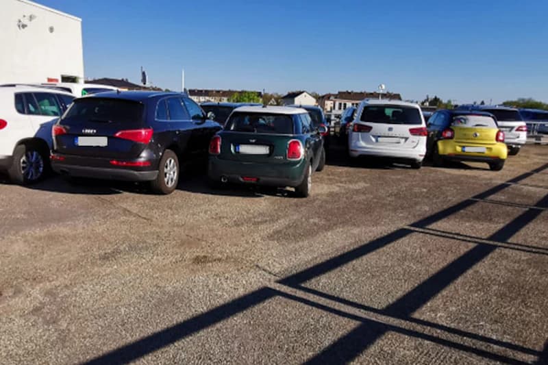 Mehrere geparkte Autos auf offenem Parkplatz mit Asphaltboden, Gebäuden im Hintergrund und langen Schatten einer Umzäunung.