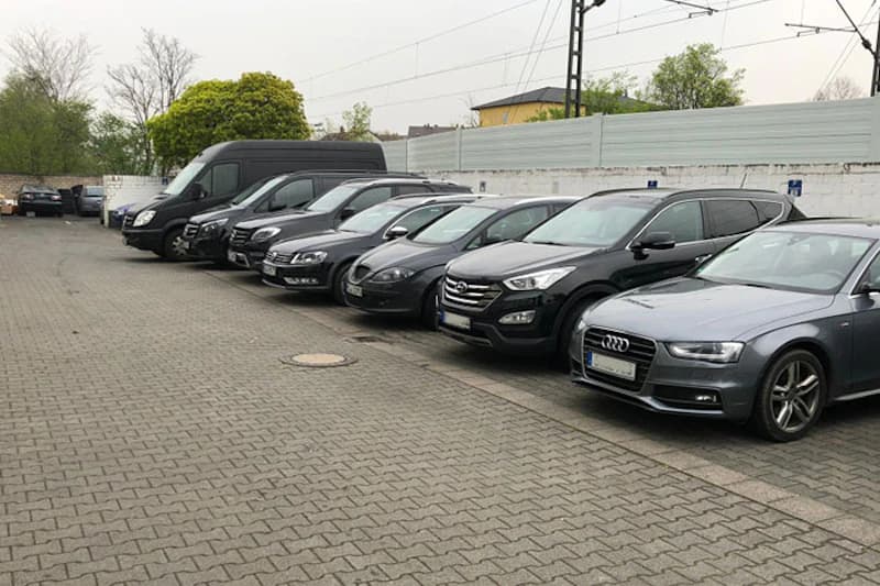Reihe geparkter Pkw und Transporter auf gepflastertem Hof vor weißer Mauer und Zaun.
