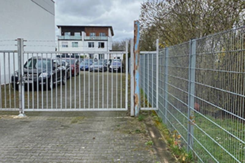 Metallgittertor vor umzäuntem Parkplatz mit mehreren Fahrzeugen und Gebäude im Hintergrund.