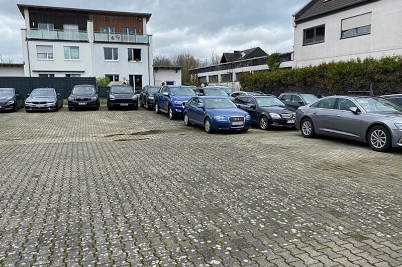 Gepflasterter Außenparkplatz mit zwei Reihen geparkter Autos vor Wohngebäuden bei bewölktem Himmel.