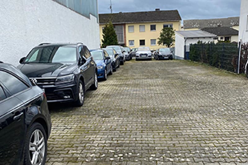 Parkanlage mit gepflastertem Boden, links nebeneinander geparkte Autos und Wohnhäuser im Hintergrund.