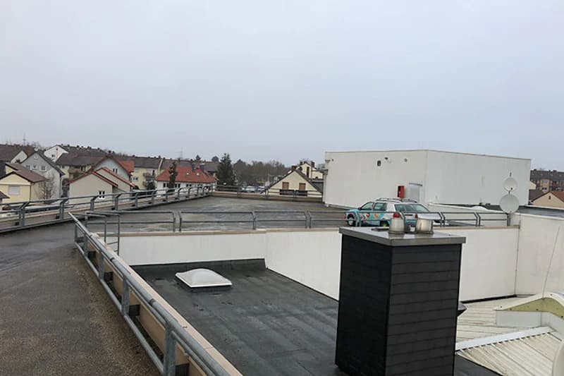 Oberes Parkdeck mit Metallgeländern, abgestelltem Pkw, Schornstein und Wohnhäusern im Hintergrund.