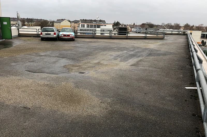 Leeres oberes Parkdeck mit nassen Pflaster, zwei geparkten Autos links und umlaufendem Metallgeländer.