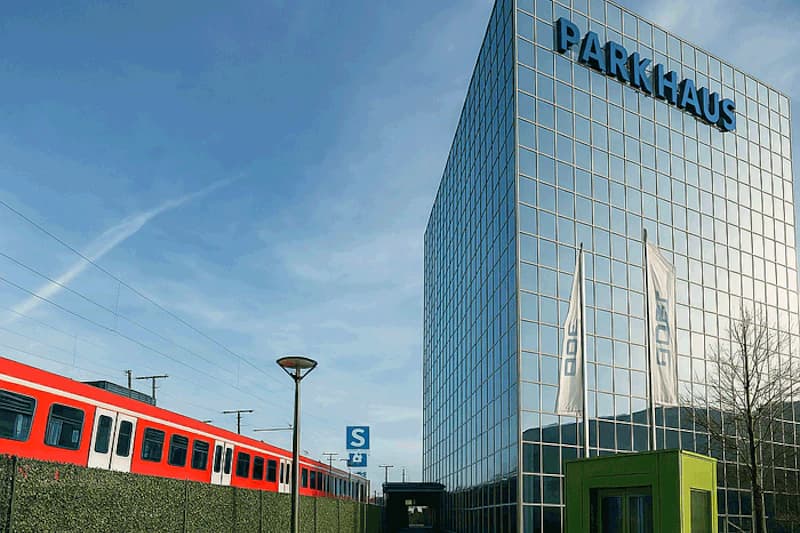 Spiegelnde Glasfassade mit PARKHAUS-Schrift, links rote S-Bahn, Fahnenmast und kleines grünes Häuschen im Vordergrund.